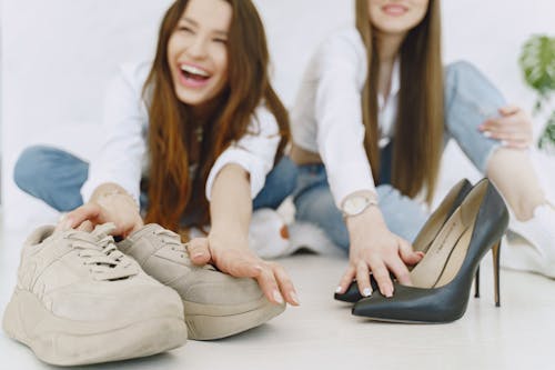 Free Crop exited young female in jeans and white shirts choosing between elegant glamour black high heels and comfortable trendy suede sneakers while sitting on floor together Stock Photo