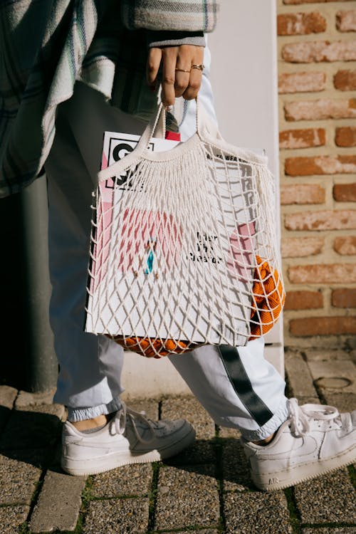 Free Crop faceless female in trendy outfit holding shopping bag with magazine while strolling on pavement Stock Photo