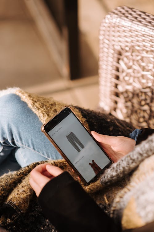 Free High angle of unrecognizable woman sitting on chair and choosing clothes in internet shop using mobile phone Stock Photo