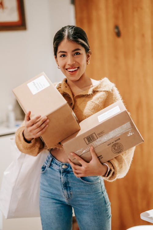 Free Smiling young ethnic lady in casual outfit standing in light room in house with packages and shopping bag while looking at camera Stock Photo